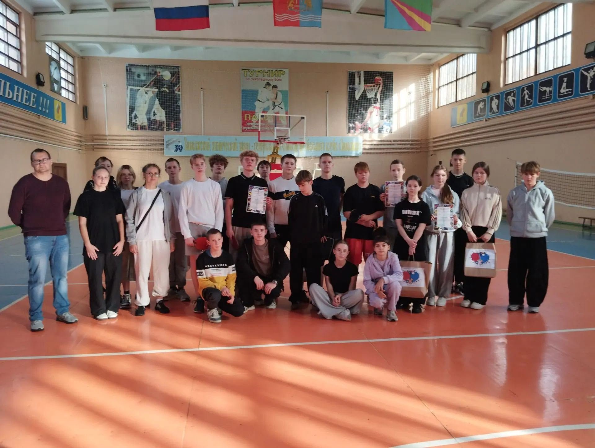 Групповая фотография школьников и взрослых в спортивном зале: участники стоят и сидят на паркете на фоне баскетбольного кольца и плаката с надписью «ТУРНИР», у некоторых — грамоты и подарочные пакеты.