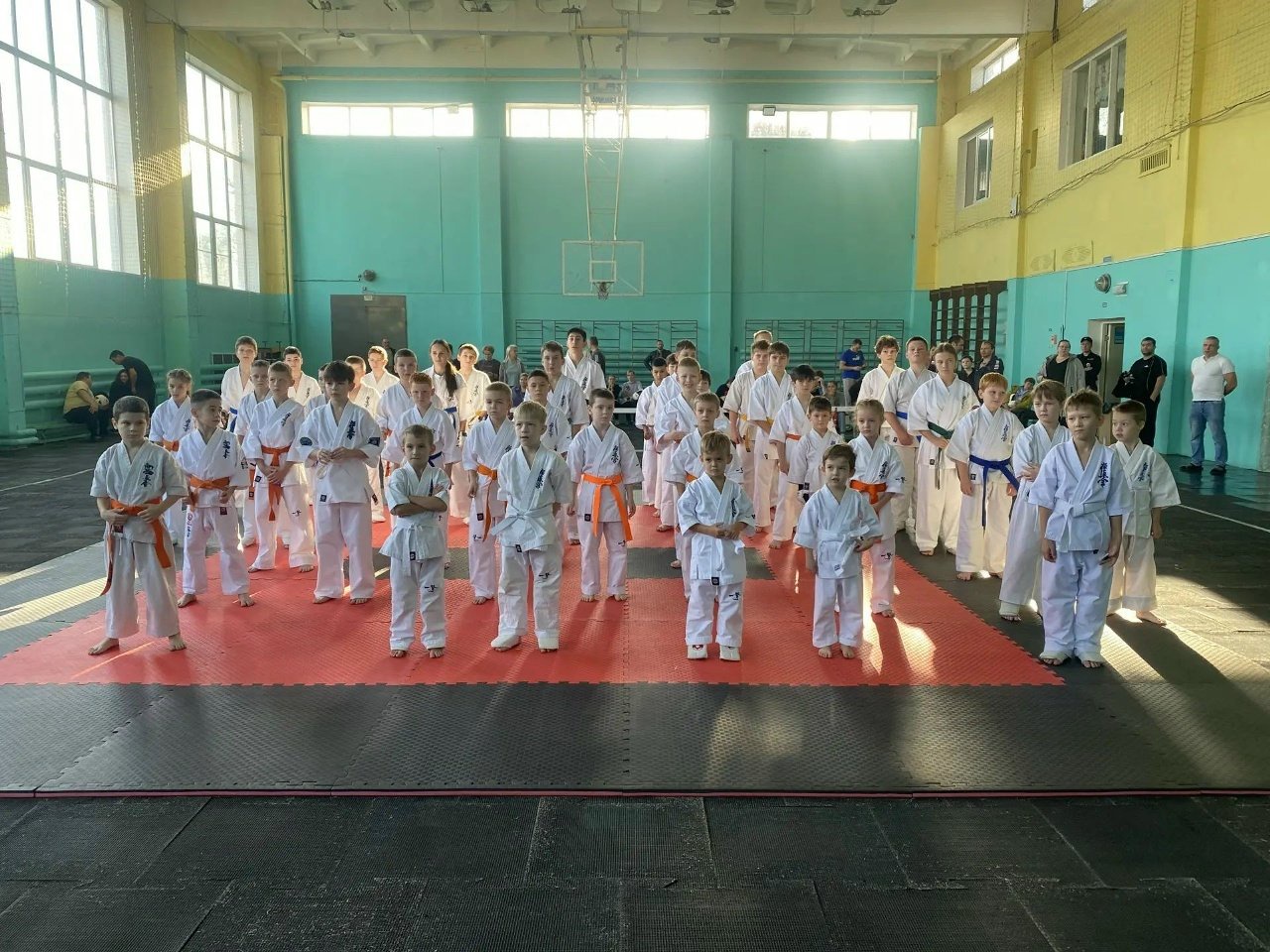 Группа юных каратистов в кимоно и поясах на красно-чёрных татами в спортивном зале